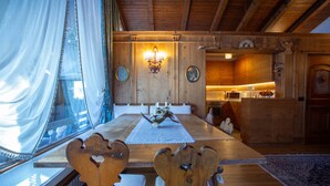 A wooden dining table with a white table runner and a decorative centerpiece. There are two wooden chairs with heart-shaped cutouts. A window with sheer curtains is visible, and a kitchen area is in the background. - Elegant Ampezzo-style apartment with breathtaking views of the Dolomites. (Cortina d'Ampezzo)
