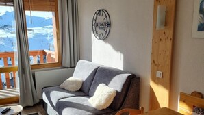 A cozy room with a sofa, a wooden table, and a view of a snowy landscape through a window. - Ski-in/ski-out & foot of the slopes, 5th floor, view ski slopes, balcony, tv, ski locker, 23m² (La Plagne)