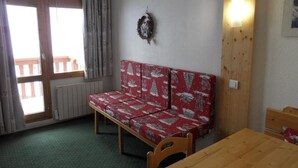 A small room with a wooden table, a bench with a red and white pattern, a radiator, and a window with curtains. - Ski-in/ski-out & foot of the slopes, 5th floor, view ski slopes, balcony, tv, ski locker, 23m² (La Plagne)