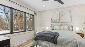 Une chambre avec une grande fenêtre, un lit recouvert d’une couverture grise et deux tableaux accrochés au mur.