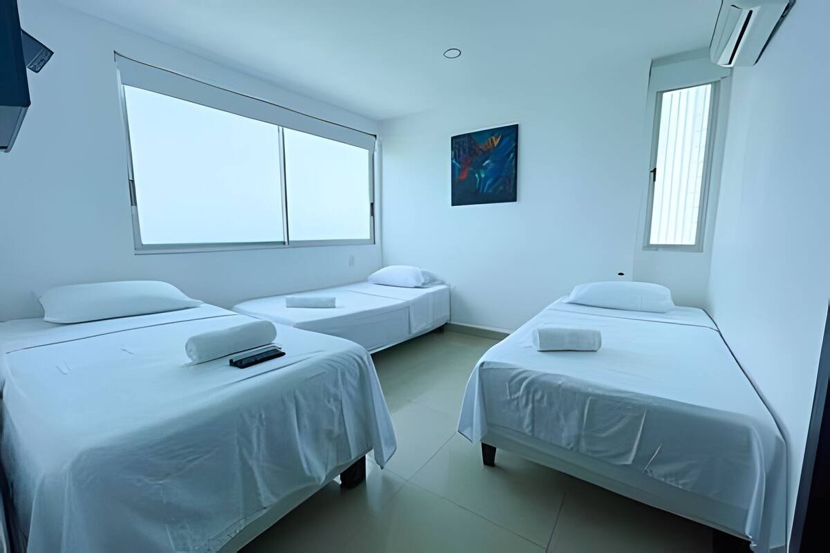 A hotel room with three single beds, a large window, an air conditioning unit, and a painting on the wall.