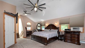 A bedroom with a large bed, a ceiling fan, a desk, a dresser, and a window with blinds.