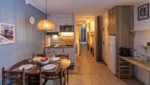A compact kitchen with a microwave, sink, and wooden cabinets. A dining table with chairs and a hanging light fixture is visible. - Le Bastan - Appt pour 4 avec Jardin (Barèges)