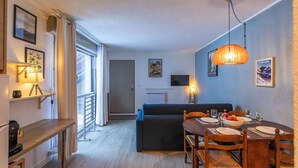 A modern living room with a sofa, dining table, and chairs. There is a TV mounted on the wall, a lamp, and a window with curtains. - Le Bastan - Appt pour 4 avec Jardin (Barèges)