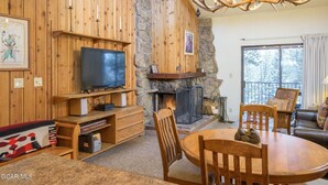A cozy living room with a stone fireplace, wooden walls, a television, and a dining area with chairs and a table. - Spacious 3Bed condo 5 min from Winter Park, walk 2 Town & steps from bus stop! (Winter Park)