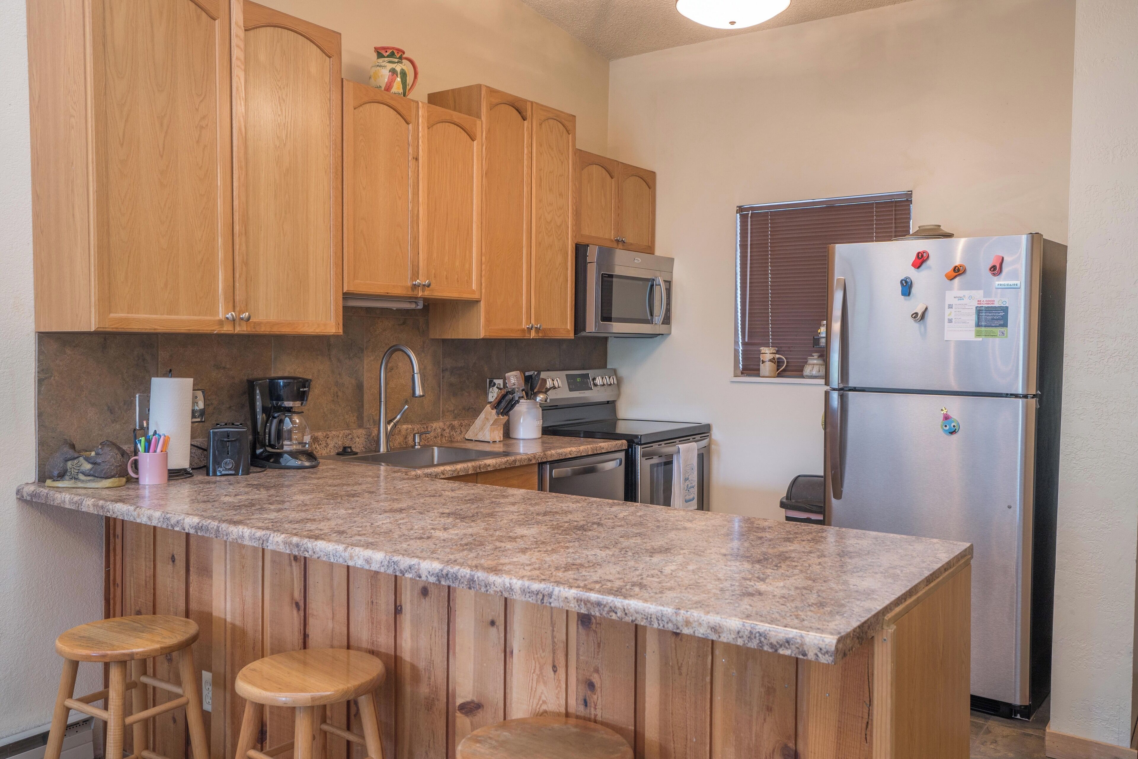 A kitchen with wooden cabinets, a granite countertop, a stainless steel refrigerator, a microwave, and a coffee maker.