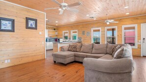 A spacious living room with a large sectional sofa, wooden walls, and a kitchen area visible in the background.