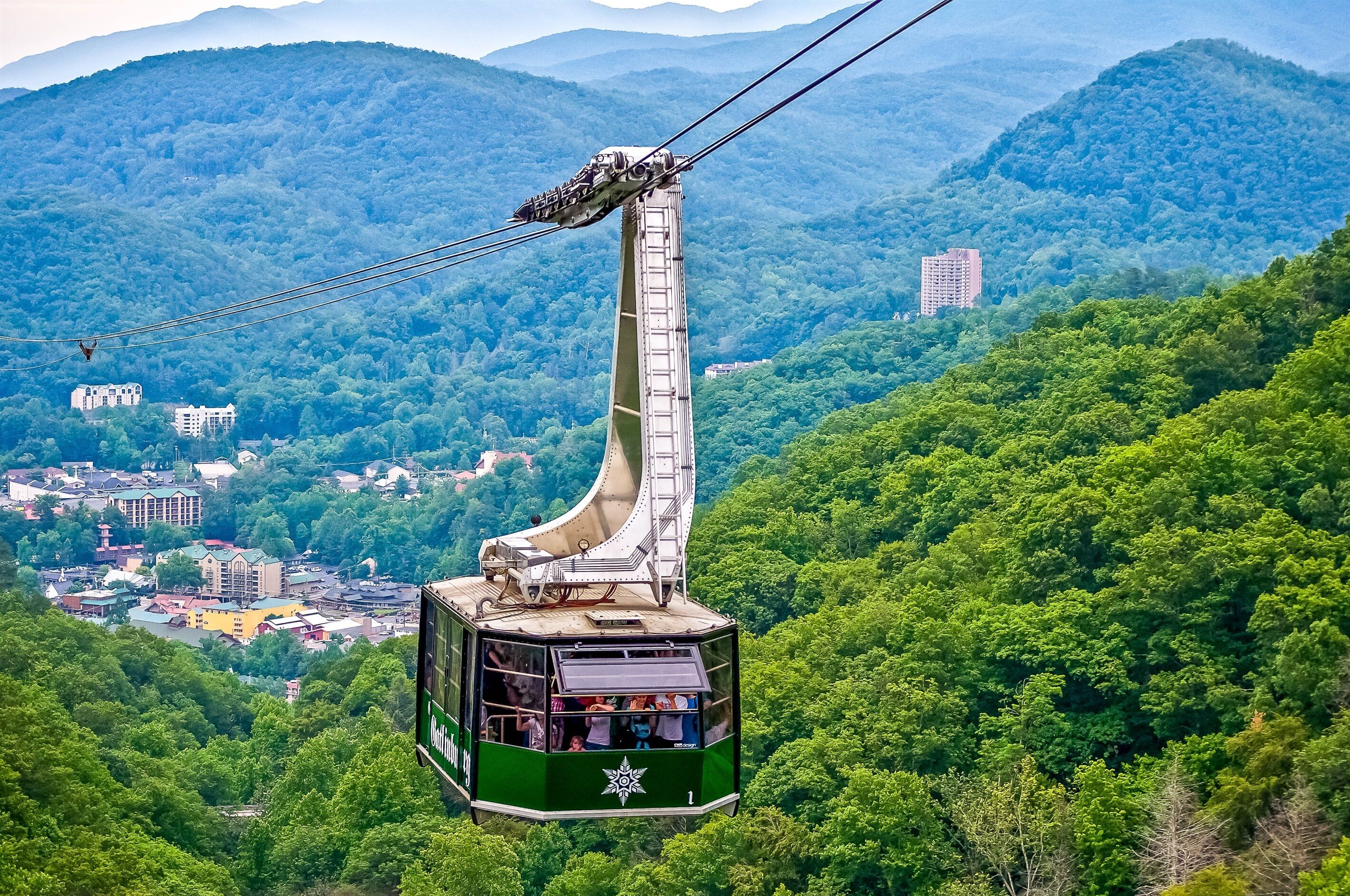 A cable car with passengers, suspended by cables, traveling over a lush green forested area with buildings in the background.