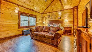 Una acogedora habitación tipo cabaña de madera, con techo de madera, un sofá de cuero marrón, una cama con cabecera blanca y un armario de madera.