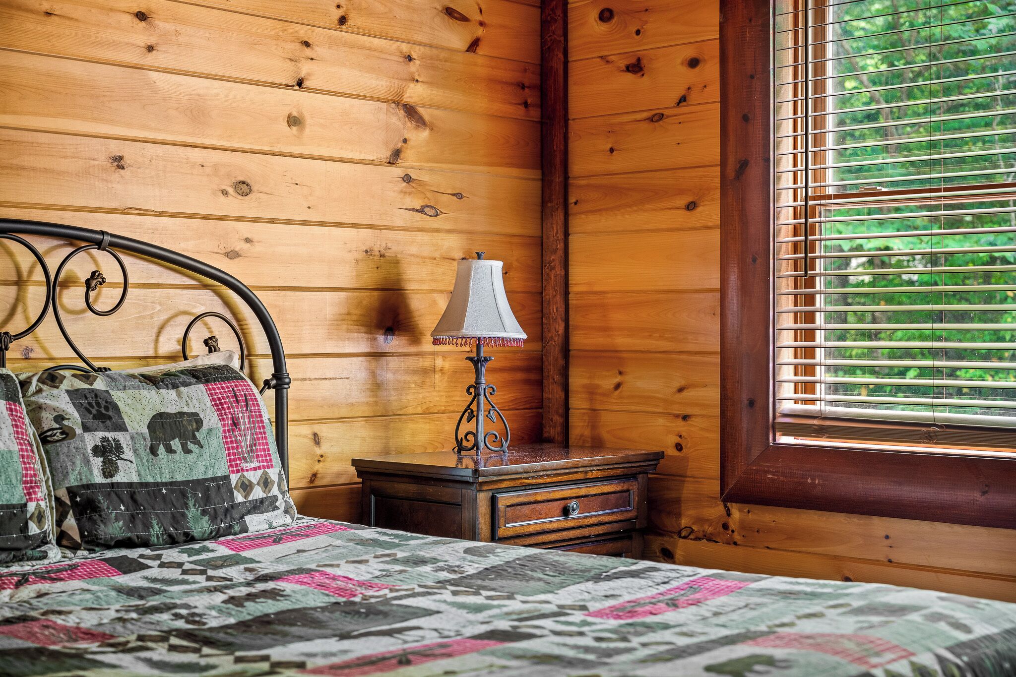 A wooden cabin room with a bed, a nightstand, a lamp, and a window with blinds.