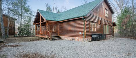 A log cabin with a green roof, wooden porch, and a gravel driveway.