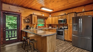 A wooden cabin kitchen with a central island, bar stools, a refrigerator, microwave, stove, and wooden cabinets. - Heated Pool, Theater, Game Room | Sleeps 25 (Cosby)