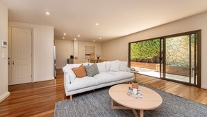 A modern living room with a light-colored sofa, a round wooden coffee table, and a large sliding glass door leading to an outdoor area with a stone wall and greenery.