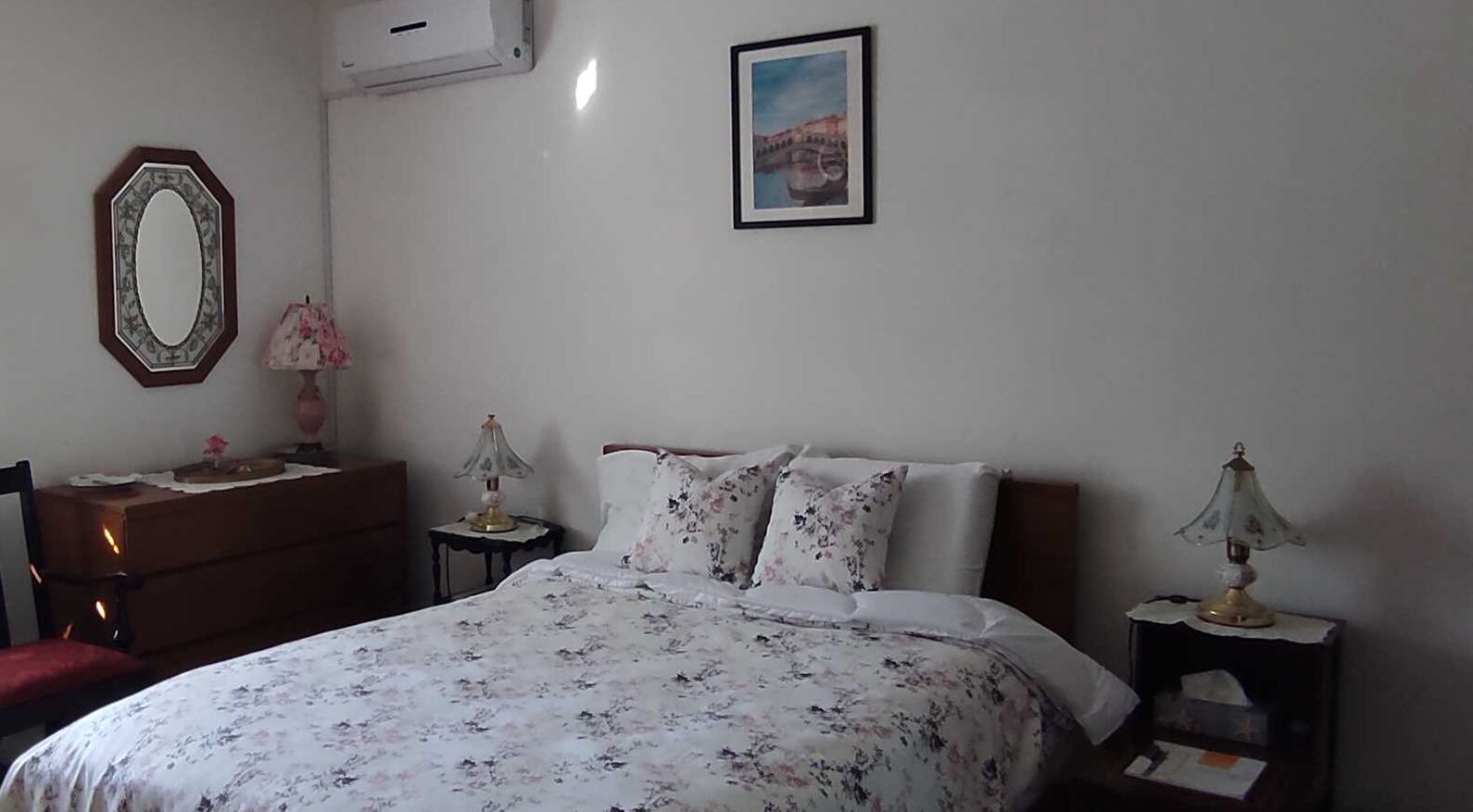 Ein Schlafzimmer mit einem Bett, einem Schminktisch, einem Spiegel, einer Lampe und einem gerahmten Bild an der Wand.