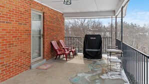 A covered balcony with red Adirondack chairs, a grill cover, and a view of trees.
