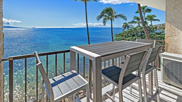 Un balcon avec une table et des chaises donnant sur une mer bleue et limpide et sur des palmiers.