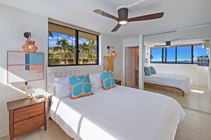 A bedroom with a large bed, a nightstand, a ceiling fan, a window with a view of palm trees, and a door leading to another room. - New Luxury Oceanfront Condo ★ Amazing 180° Ocean Views ★ Top-Floor Corner-Unit (Wailuku)