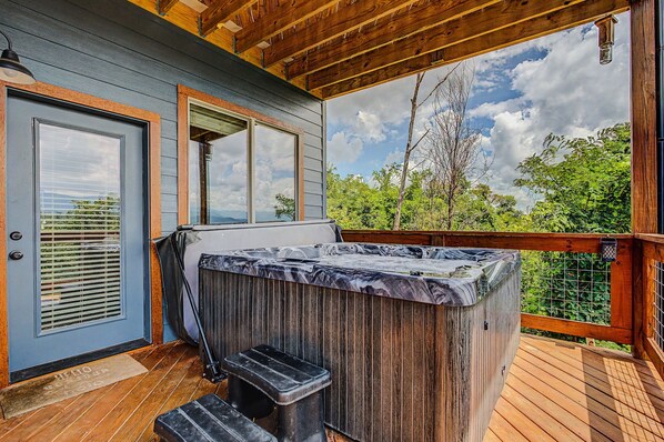 A covered outdoor deck with a hot tub, wooden deck flooring, a door with a window, and a view of trees and sky.