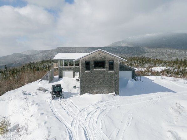 A two-story house with a garage, surrounded by snow-covered landscape and mountains.