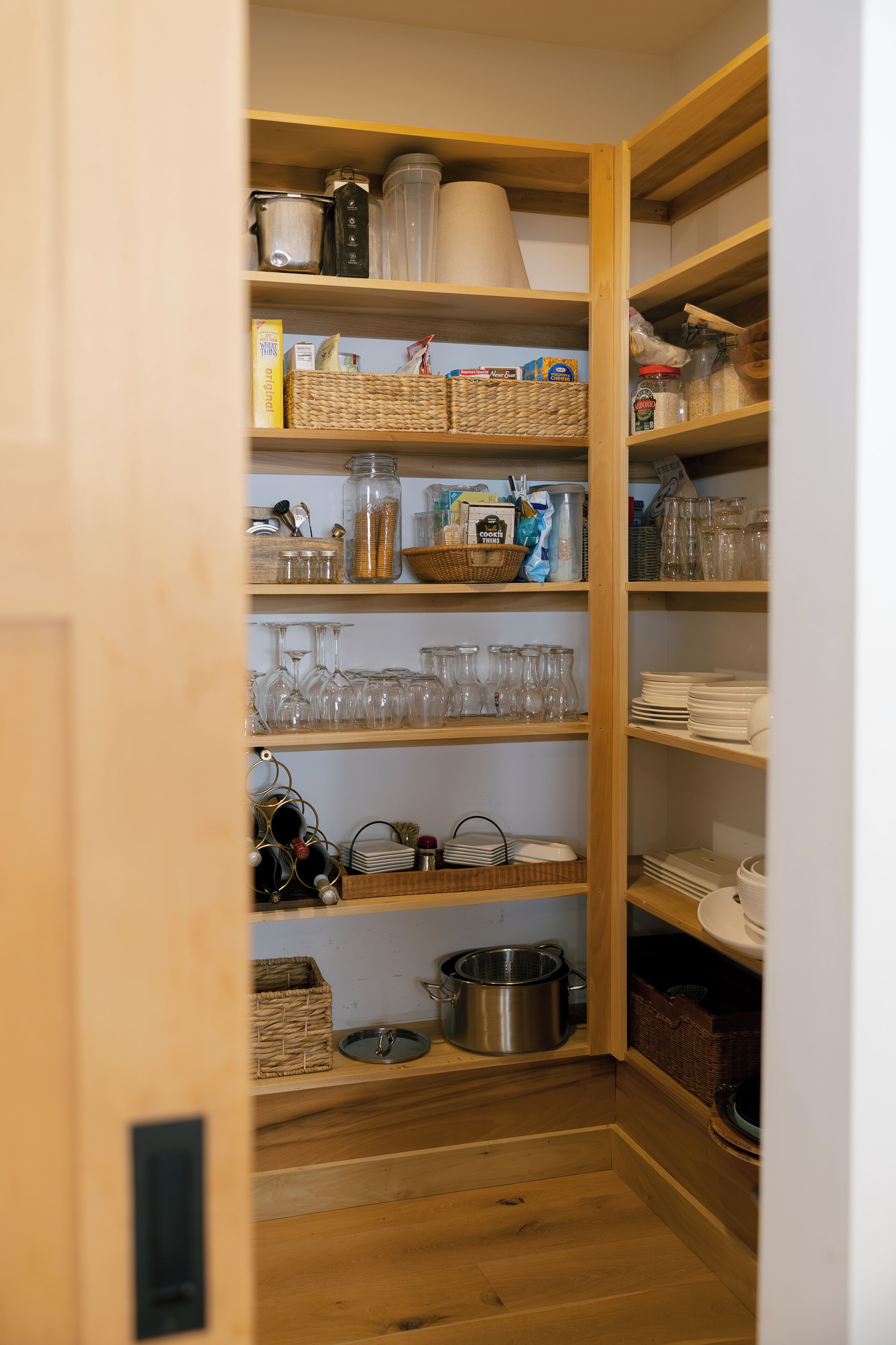 A wooden pantry with shelves containing glassware, jars, and kitchen utensils.