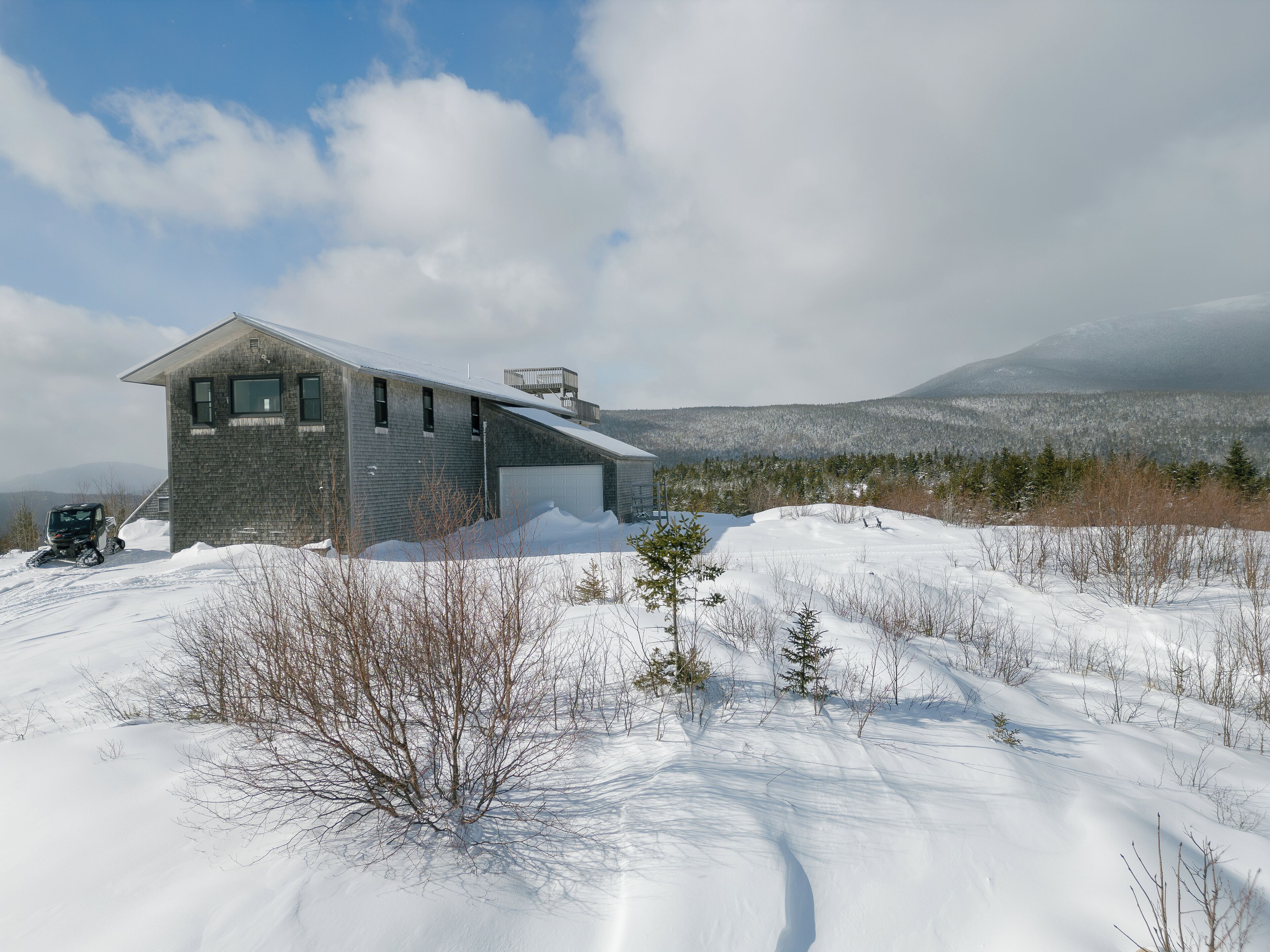 Et moderne hus med skråt tag, garage og et sneklædt landskab med bjerge i baggrunden.