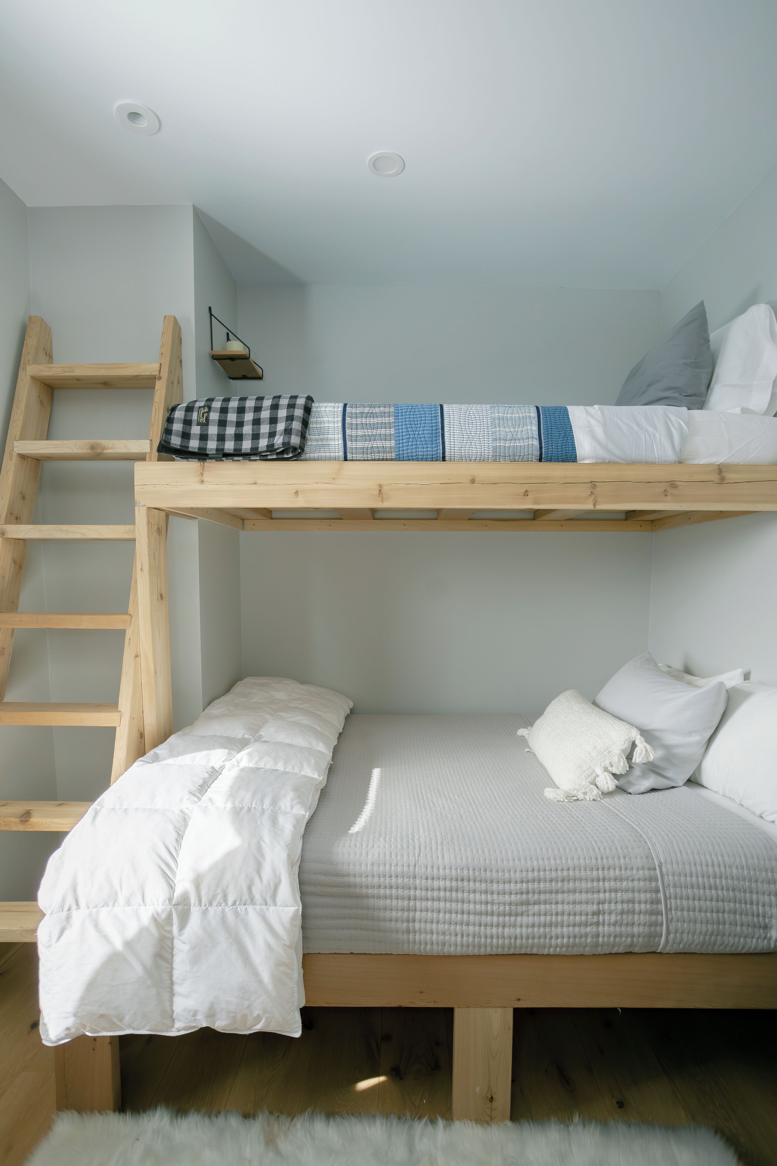 Una cama litera con escalera de madera, un pequeño estante con lámpara y una alfombra blanca y mullida.