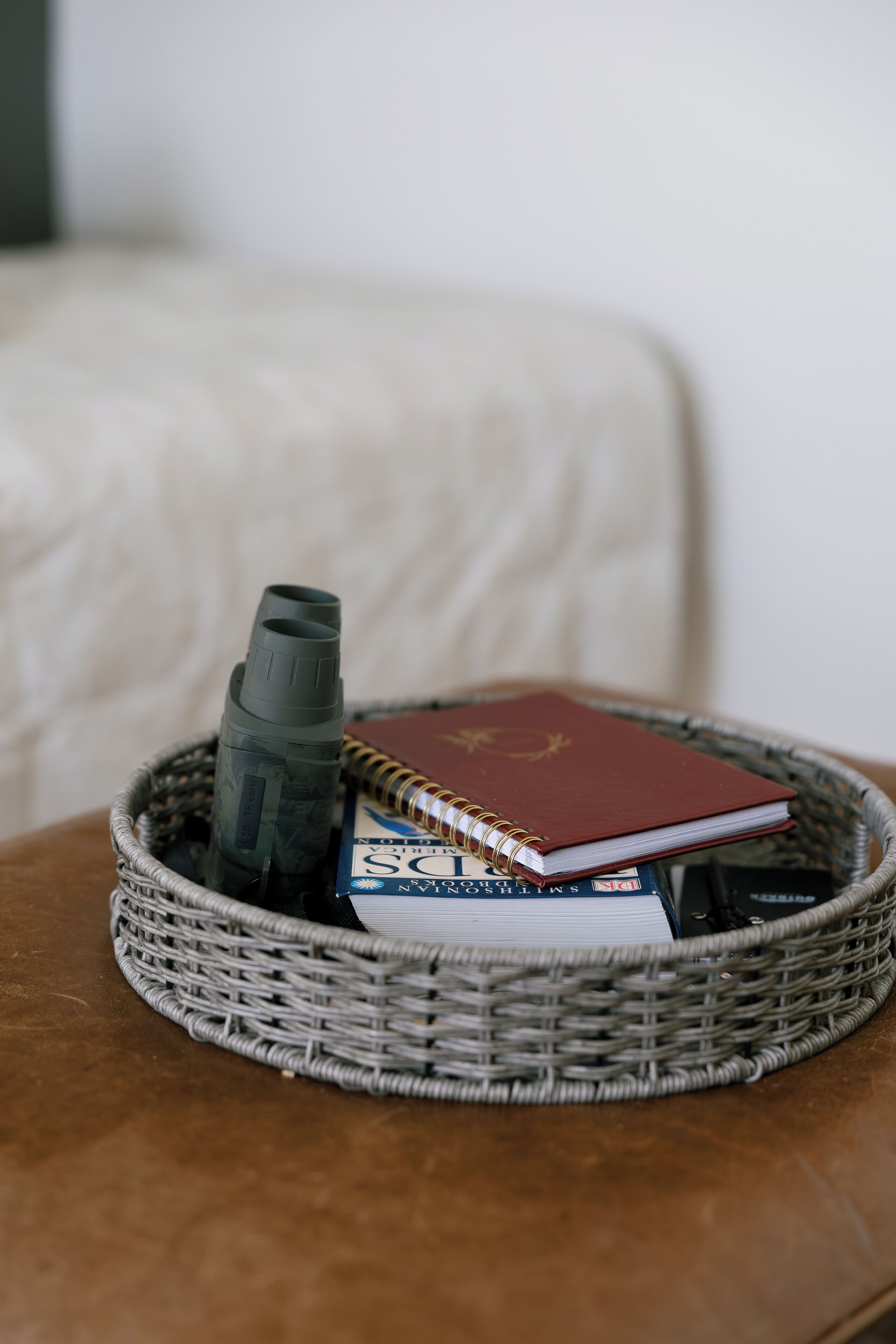A wicker basket containing a telescope, a red book, and a notebook on a brown leather ottoman.