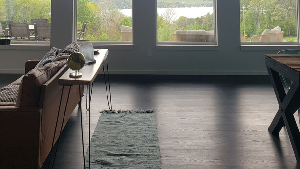 A room with large windows offering a view of a lake and surrounding greenery. There is a wooden bench, a small table, and a sofa.