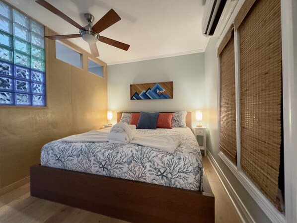 Une chambre à coucher comprenant un lit, un ventilateur de plafond, une fenêtre avec des stores et une œuvre d’art accrochée au mur.