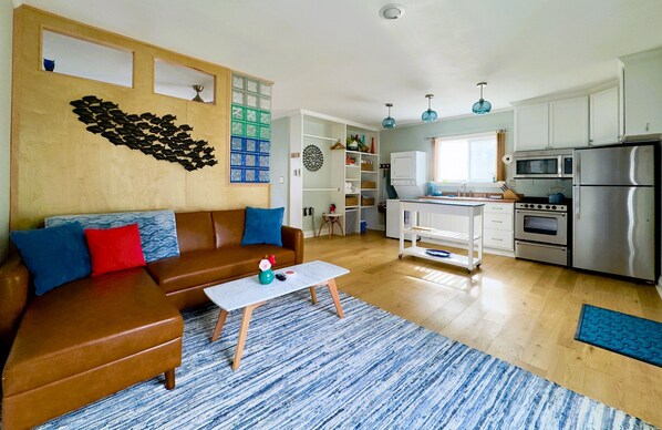 A modern living room with a brown leather sofa, a coffee table, and a blue patterned rug. The kitchen features white cabinets, stainless steel appliances, and a blue hanging light fixture.