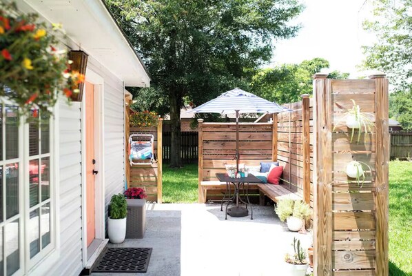 Un patio con un banco de madera, una mesa y un sillón colgante. Hay un jardín pequeño con plantas en macetas y una cerca de madera al fondo.