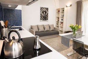 Eine moderne Küche mit Wasserkocher, Kaffeemaschine und Spüle. Ein graues Sofa mit Kissen, ein kleiner Tisch mit einem Laptop und eine Vase mit gelben Blumen.