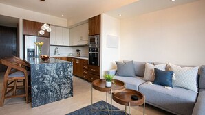 Una cucina moderna con isola in marmo, mobili in legno ed elettrodomestici in acciaio inossidabile. Si nota un divano grigio con cuscini decorati e un piccolo tavolino in legno con una pianta.
