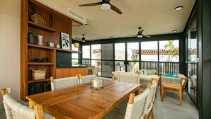 Un'ampia sala da pranzo con tavolo in legno e sedie in vimini, una libreria in legno e un balcone con vista su palme.