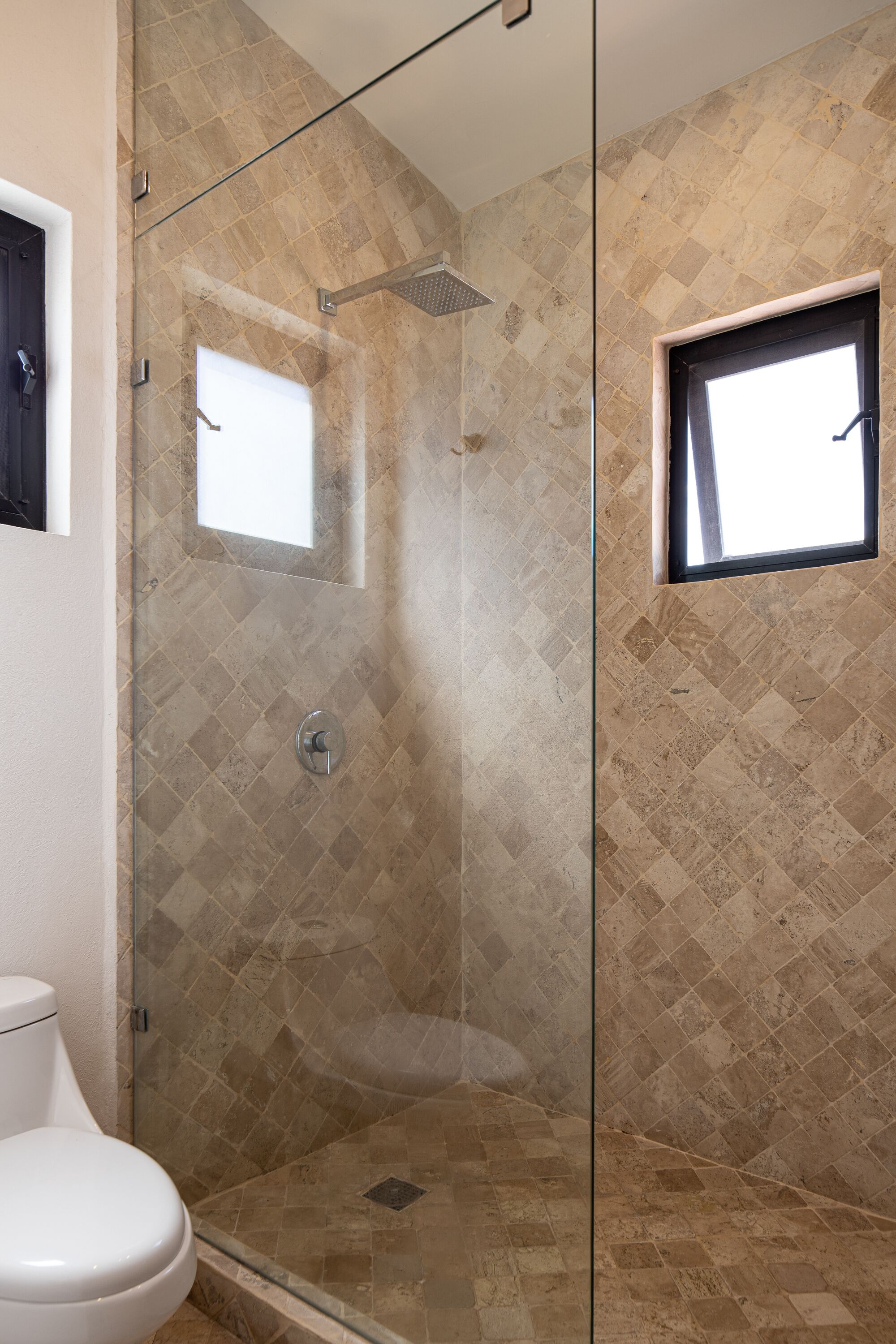 A glass-enclosed shower with a tiled wall, a window, and a toilet visible to the left.