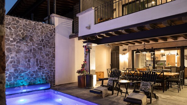 Un área de piscina moderna al aire libre con paredes de piedra, una terraza de madera y un comedor con mobiliario exterior.