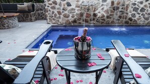 Zona junto a la piscina con pared de piedra, sillones de descanso, mesa con cubo para champán y fuente de agua.