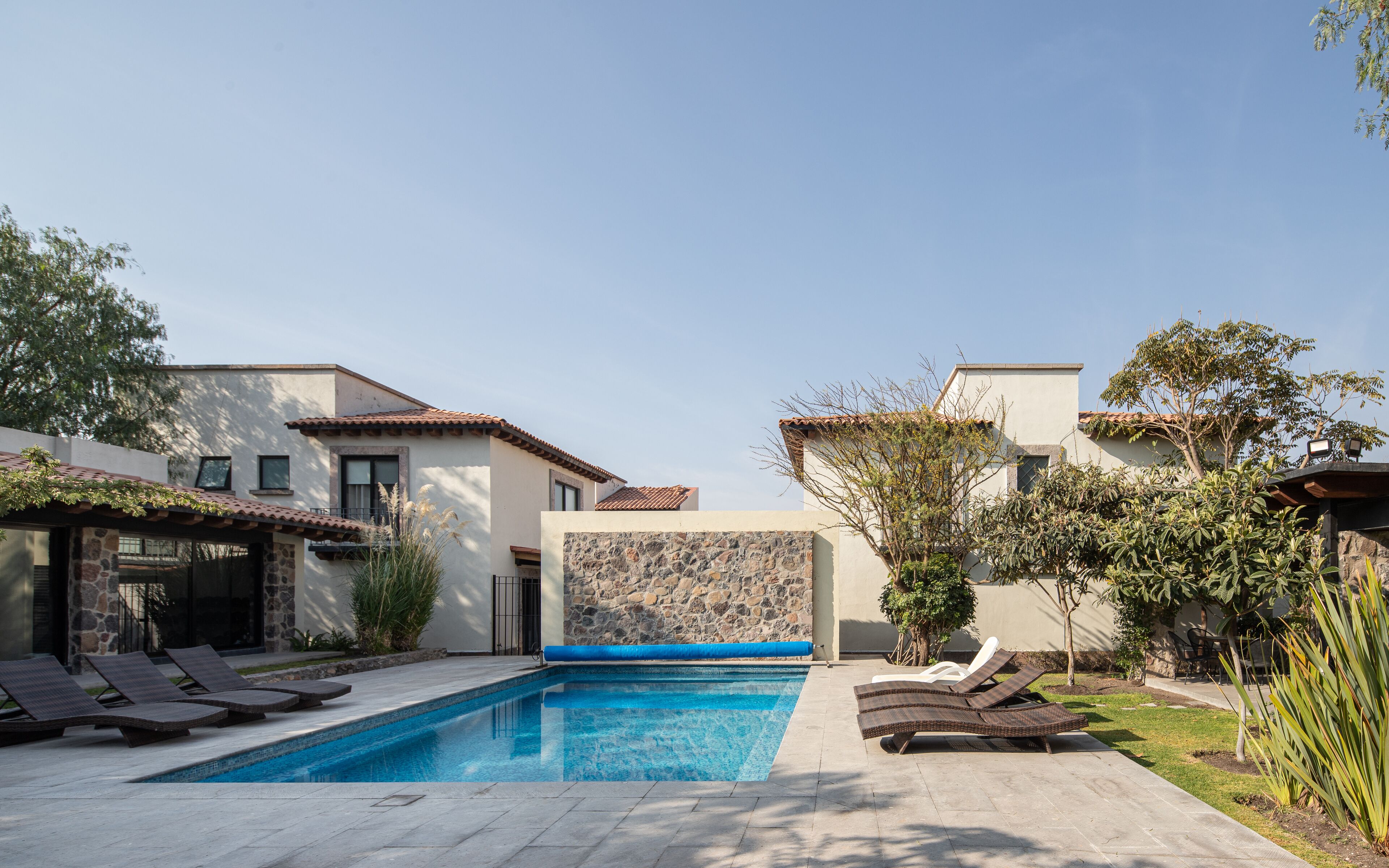 A swimming pool with lounge chairs, a stone wall, and two houses with tiled roofs.