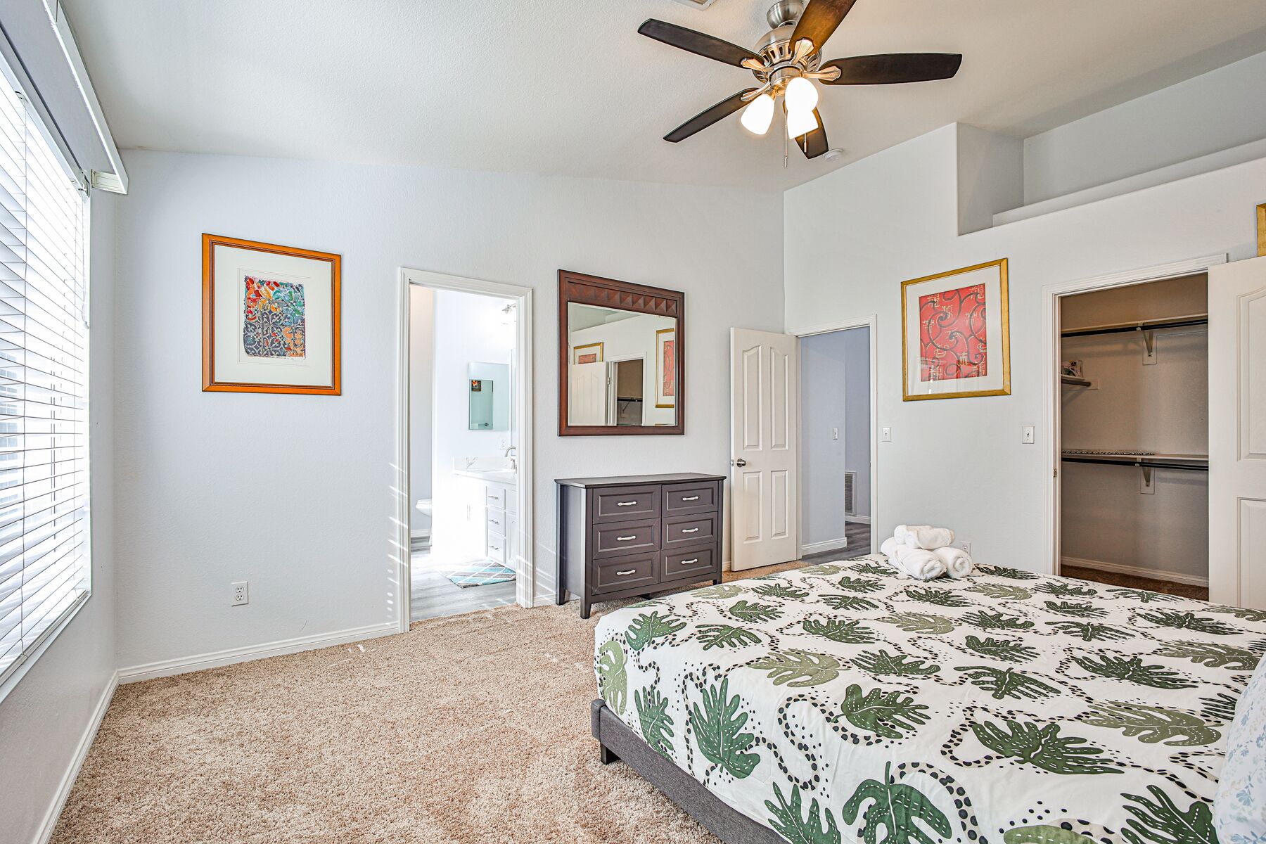 A bedroom with a bed, a ceiling fan, a dresser, a mirror, and a door leading to a bathroom.