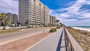 Un hotel de varios pisos con un paseo pavimentado a lo largo de la playa, cielo despejado y palmeras.