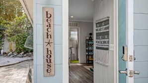 A wooden sign reading "beach house" on a wall, a doorway leading to a room with a patterned rug, and a shelf with various items.