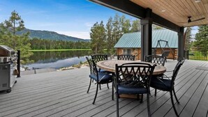 Eine überdachte Terrasse mit rundem Tisch und Stühlen, mit Blick auf einen See und bewaldete Hügel.