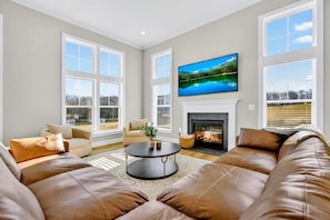 A living room with a fireplace, large windows, a television, and a leather sofa. - Lakefront Luxury - NEW 6BR, Lake Anna, Dock, Games (Mineral)