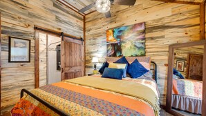 A bedroom with a wooden ceiling, a bed with colorful bedding, a ceiling fan, and a large abstract painting on the wall.