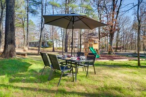 Een buitenterras met een tafel en stoelen onder een grote parasol, omgeven door bomen en een speeltuin.