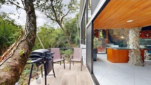 An outdoor patio with a grill, seating, and a view of a restaurant area with stone pillars and a wooden ceiling. - Artsy Riverfront Jungle Retreat: River Pool & Hot Tub in living room (Rivas)