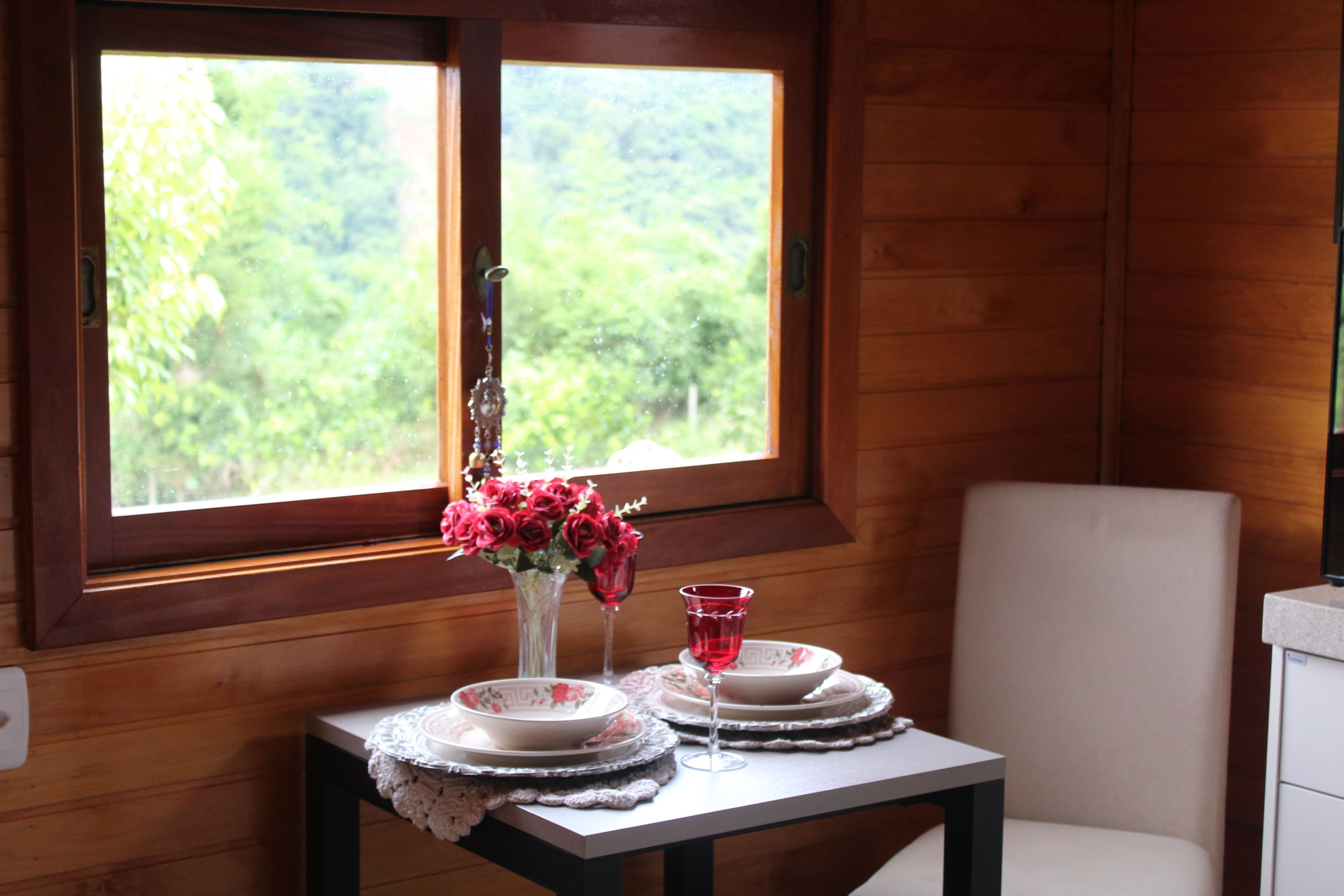 Un interior de madera con una mesa preparada para dos, un jarrón con flores rojas y una ventana con vistas a follaje.