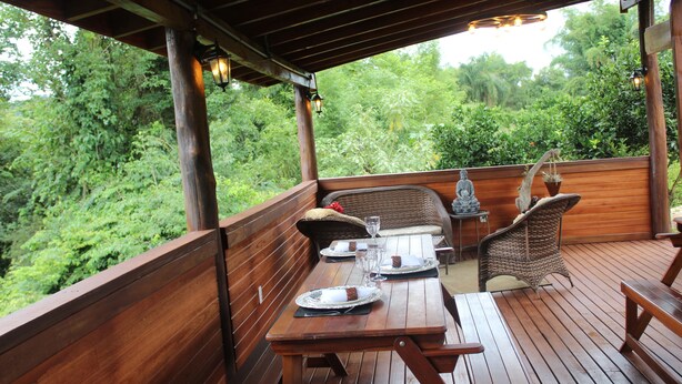 Eine Holzterrasse mit Tisch für zwei, umgeben von Korbstühlen, mit Blick auf einen üppigen, grünen Wald.