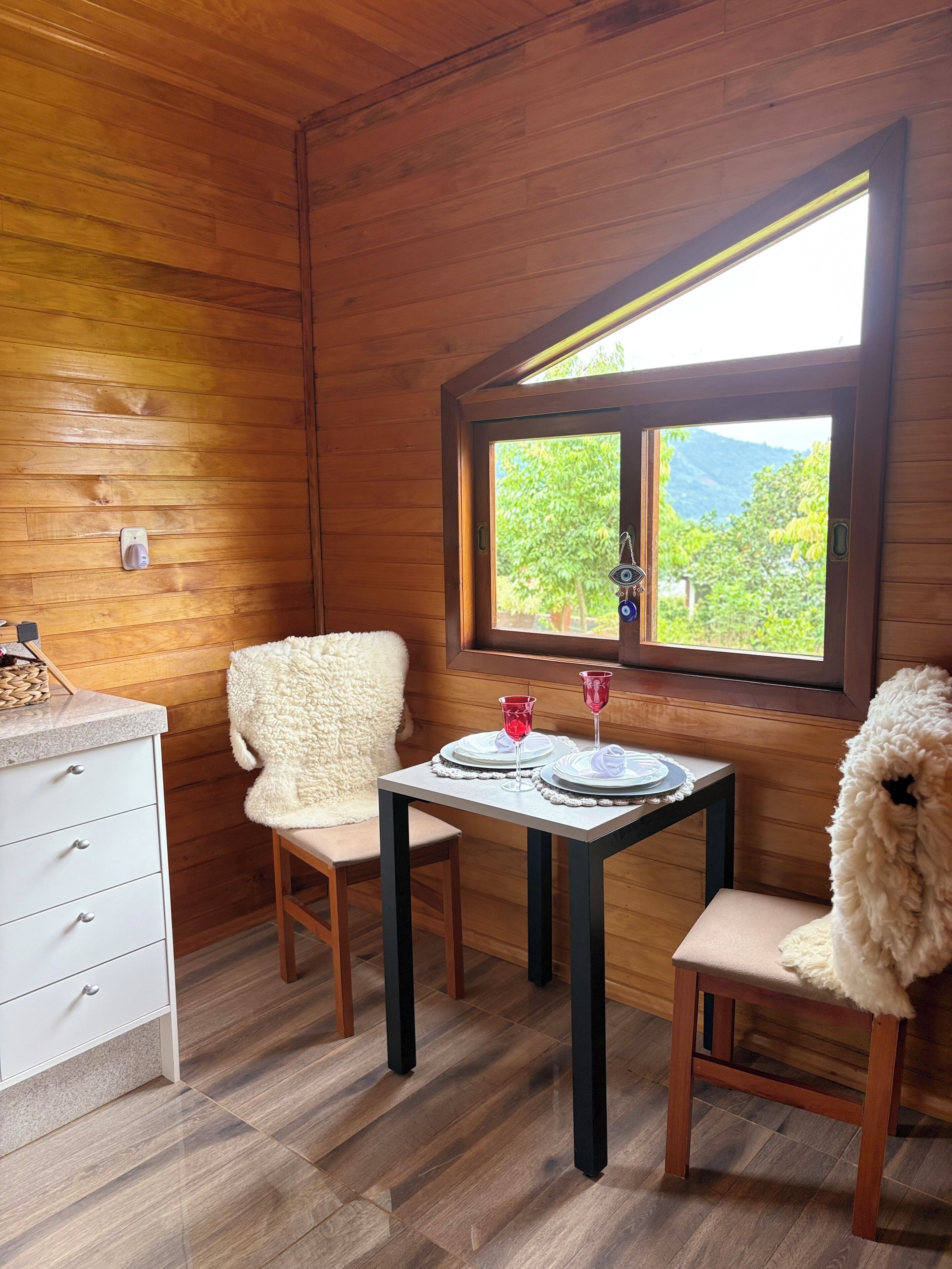 L’intérieur chaleureux d’un chalet, avec un décor en bois, une table à manger dressée avec des assiettes et des verres, et une fenêtre donnant sur un paysage verdoyant.