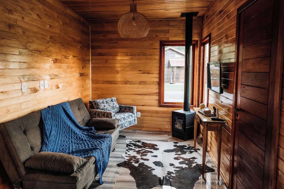 Ein gemütliches Blockhaus-Interieur mit einer Couch, einer Decke, einem Kuhfellteppich, einem Kaminofen und einem Fenster mit Blick auf ein Gebäude.
